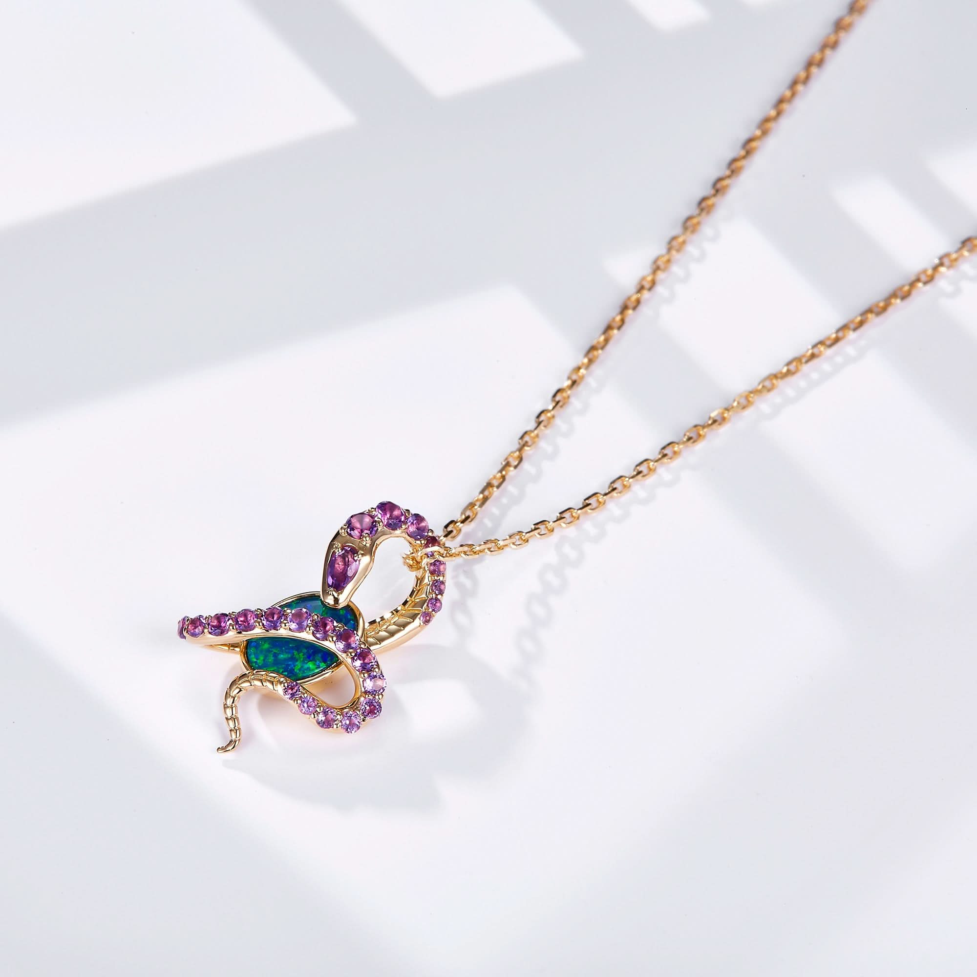 An 18K gold pendant necklace featuring an oval Australian opal wrapped in an intricate snake design adorned with natural amethyst gemstones. The opal displays vibrant blue-green hues, beautifully contrasted by the rich purple of the amethysts and the warm yellow gold setting. The pendant hangs from a delicate gold chain, creating a bold yet elegant statement piece.
