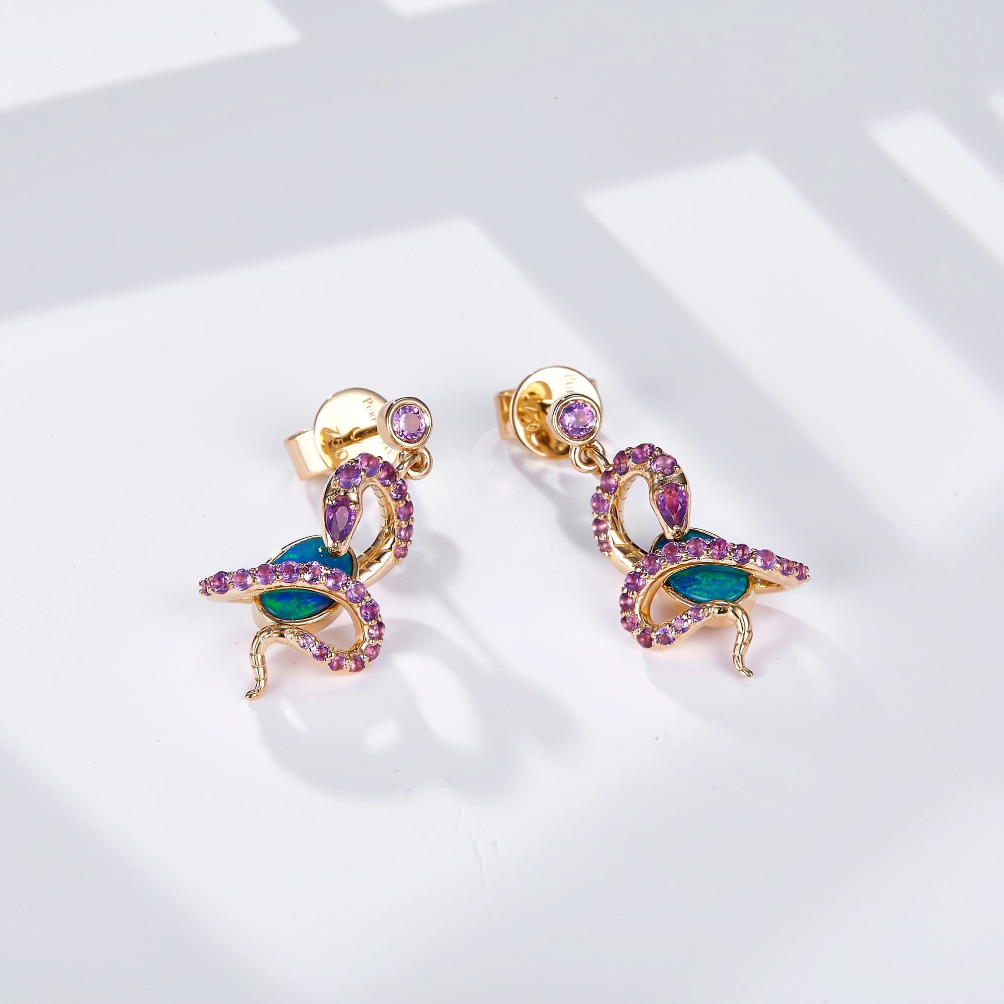 A pair of 18K gold dangle earrings featuring an intricate snake design wrapped around oval Australian opals, adorned with natural amethyst gemstones for a striking contrast. A round amethyst gemstone accents the stud, enhancing the luxurious appeal. The opals display vibrant blue-green hues, beautifully set in yellow gold. These statement earrings blend bold artistry with elegant sophistication, perfect for unique and luxurious styling.