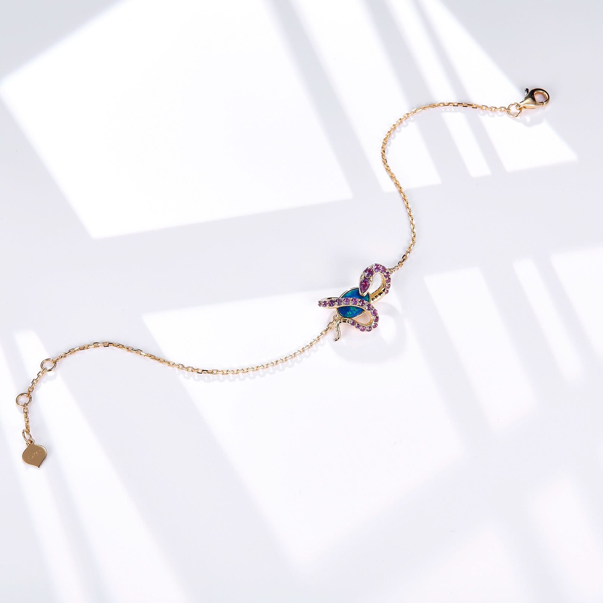 An elegant 18K gold bracelet featuring a snake design wrapped around an oval Australian opal. The snake is adorned with sparkling amethyst gemstones, complementing the vibrant blue-green hues of the opal. The delicate gold chain is secured with a lobster clasp and includes a small heart-shaped tag for a refined touch. This bracelet blends luxury with unique design, perfect for statement styling.