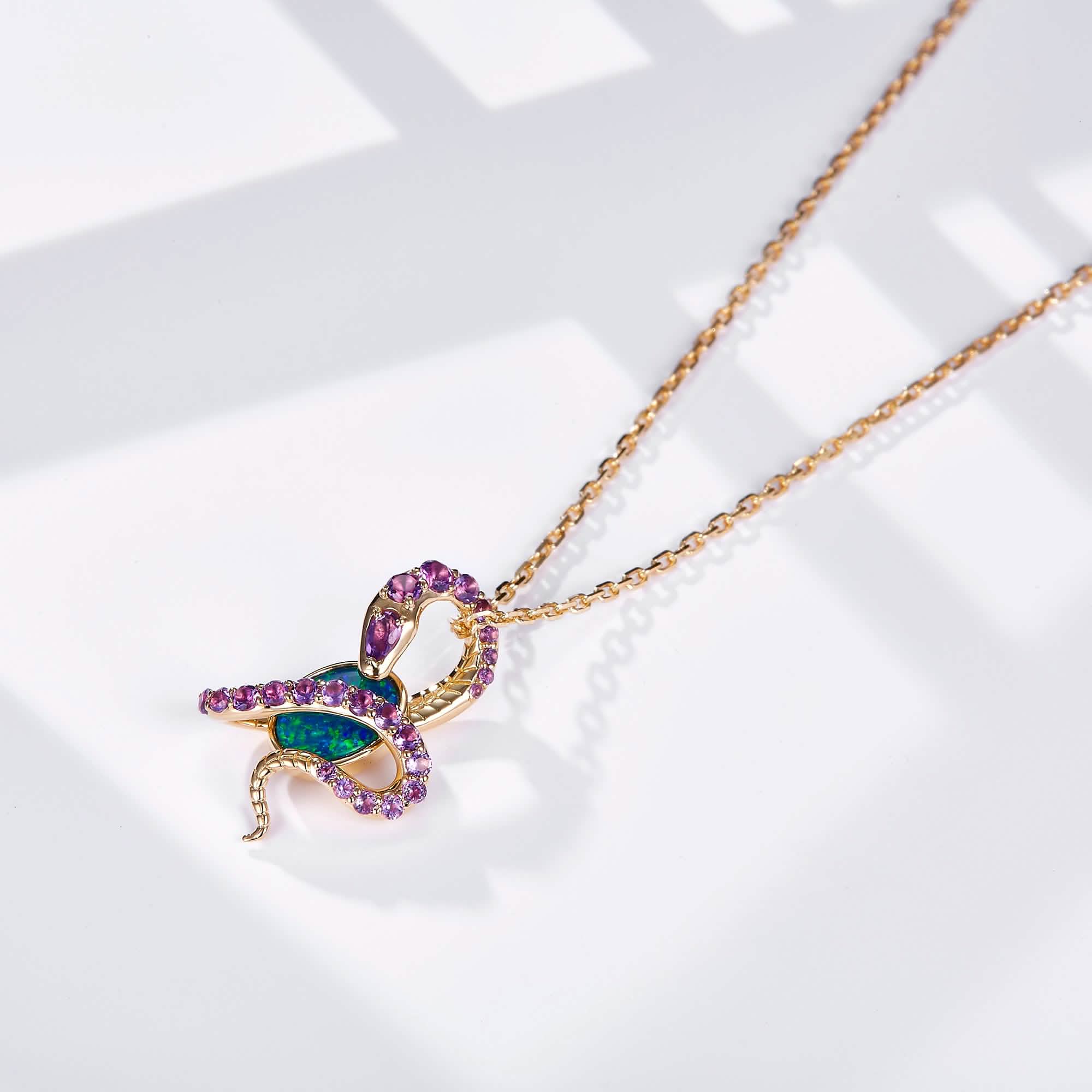 An 18K gold pendant necklace featuring an oval Australian opal wrapped in an intricate snake design adorned with natural amethyst gemstones. The opal displays vibrant blue-green hues, beautifully contrasted by the rich purple of the amethysts and the warm yellow gold setting. The pendant hangs from a delicate gold chain, creating a bold yet elegant statement piece.