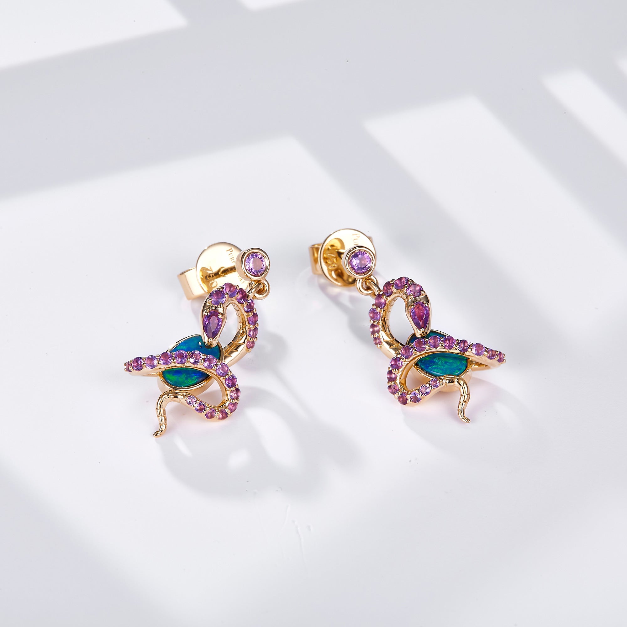 A pair of 18K gold dangle earrings featuring an intricate snake design wrapped around oval Australian opals, adorned with natural amethyst gemstones for a striking contrast. A round amethyst gemstone accents the stud, enhancing the luxurious appeal. The opals display vibrant blue-green hues, beautifully set in yellow gold. These statement earrings blend bold artistry with elegant sophistication, perfect for unique and luxurious styling.