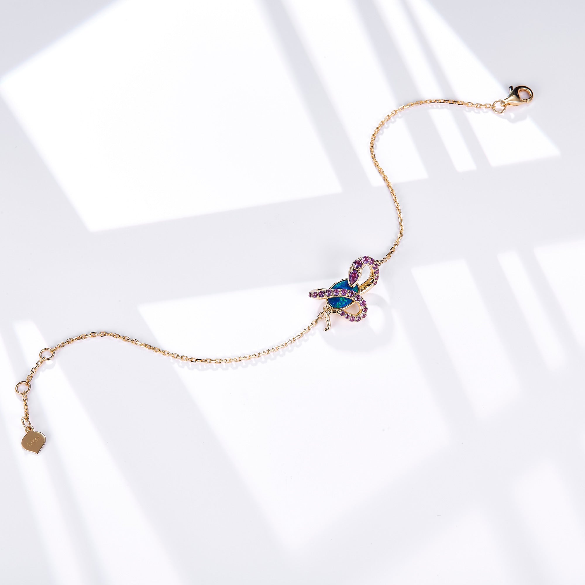 An elegant 18K gold bracelet featuring a snake design wrapped around an oval Australian opal. The snake is adorned with sparkling amethyst gemstones, complementing the vibrant blue-green hues of the opal. The delicate gold chain is secured with a lobster clasp and includes a small heart-shaped tag for a refined touch. This bracelet blends luxury with unique design, perfect for statement styling.