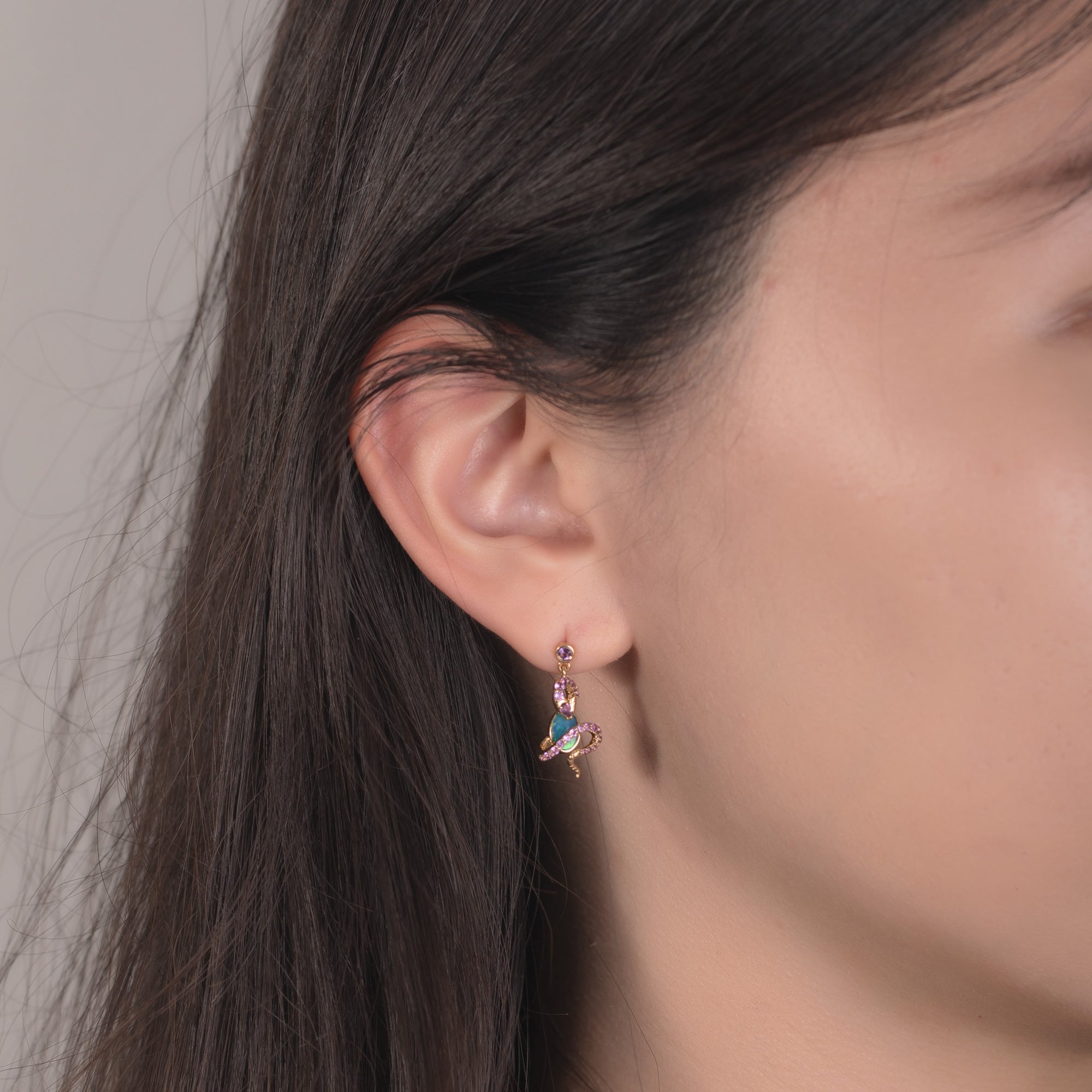 A close-up of a model wearing an 18K gold Australian opal dangle earring featuring a snake design. The earring showcases a vibrant oval Australian opal wrapped by a gold snake adorned with natural amethyst gemstones, with a round amethyst gemstone stud at the top. The opal’s blue-green hues contrast beautifully with the yellow gold and rich purple accents, creating a striking and elegant look.